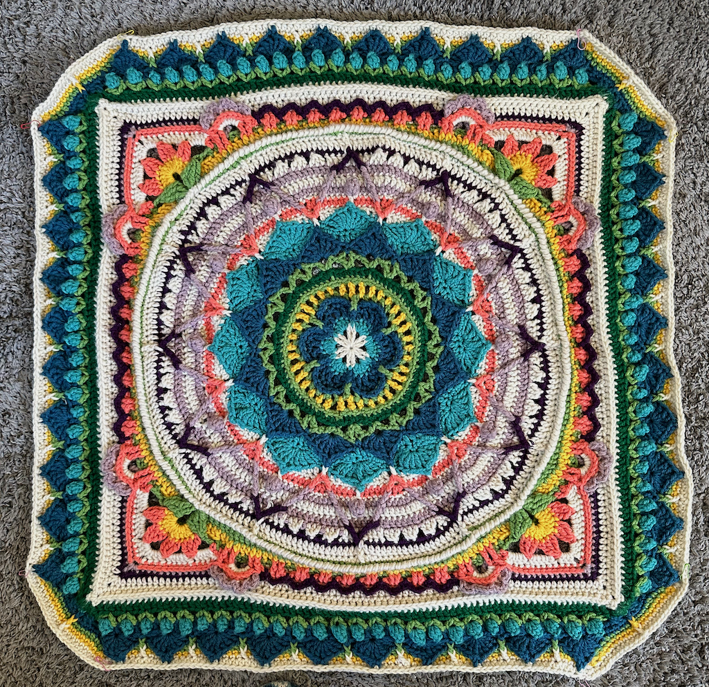 A partially completed, very intricate crochet blanket. The shape starts in the middle as a six-petaled flower and gradually becomes a circle, then a square (pink flowers form the corners), then the corners become another 4 sides, making it a (very square-looking) octagon. The colors are two blues, two greens, yellow, white, purple, and pink.
